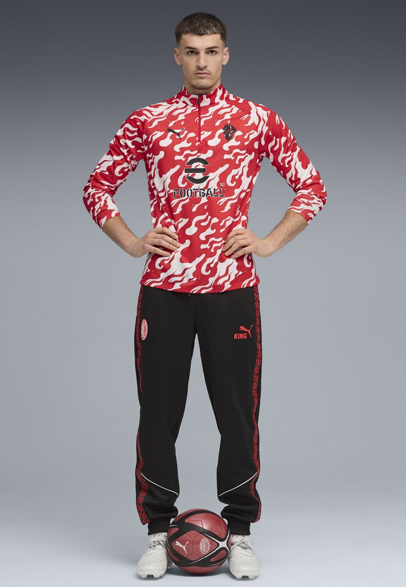 Röd och vit camo långärmad topp med svarta byxor som har röda accenter. Inkluderar logotypdesigner och en fotboll vid modellens fötter.