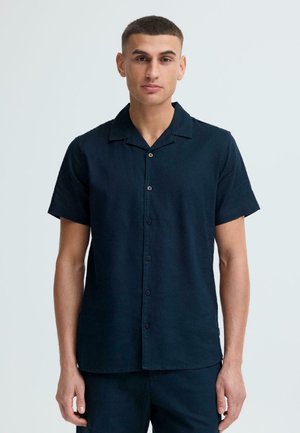 Jeune homme aux cheveux courts portant une chemise bleu marine sombre à manches courtes avec des boutons, sur un fond clair uni.