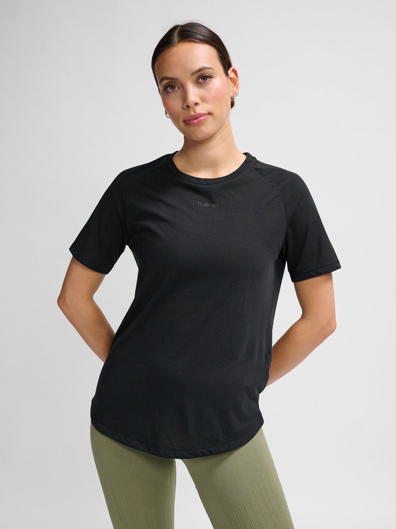 Jonge vrouw met donker haar draagt een zwart T-shirt met korte mouwen en olijfgroene broek, staand met handen achter de rug tegen een effen achtergrond.