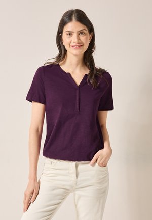 Kurzärmliges, dunkelviolettes T-Shirt mit V-Ausschnitt und einem Knopf-Detail, kombiniert mit hellen Hosen. Glatte Textur.