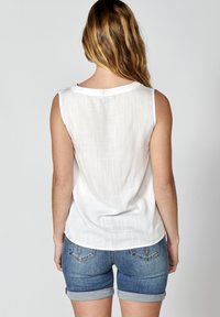 Top bianco senza maniche in tessuto leggero e testurizzato con orlo curvo. Indossato con pantaloni corti di jeans blu con risvolti e tasche sul retro.