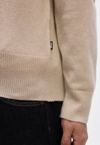 Μπεζ πλεκτό πουλόβερ με ριμπ τελείωμα και μανσέτες, φορεμένο με σκούρο τζιν, παρουσιάζοντας μια μικρή μαύρη ετικέτα στη πλευρά με την επιγρα φή "BOSS".