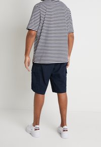 Muž stojící zády kameře, oblečený v černobílem pruhovaném tričku, námořnických cargo šortkách a bílých teniskách na jednobarevném pozadí.