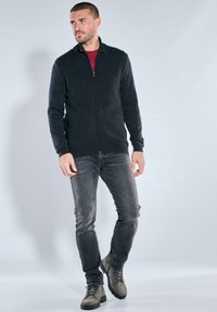 Schwarze Reißverschlussjacke aus strukturiertem Stoff mit zwei Seitentaschen, getragen über einem roten Hemd, kombiniert mit grauer Jeans und grünen Stiefeln.