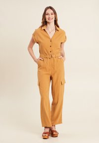 Mosterdkleurige jumpsuit met een kraag, korte mouwen, knopen aan de voorkant en een ceintuur in de taille. Inclusief zijzakken en een recht gesneden ontwerp.