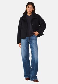 Giacca in misto lana scura con colletto in shearling, abbinata a un maglione scuro e jeans a zampa d'elefante blu, caratterizzati da una vestibilità comoda e dettagli con tasche.