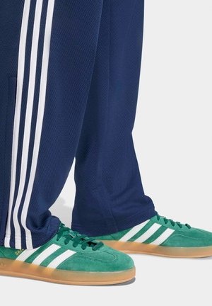 Sneakers verdi Adidas Gazelle con strisce bianche indossate con pantaloni della tuta blu navy con strisce bianche laterali su uno sfondo chiaro.