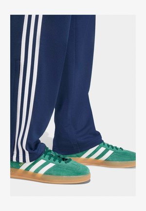Sneakers verdi Adidas Gazelle con strisce bianche indossate con pantaloni della tuta blu navy con strisce bianche laterali su uno sfondo chiaro.