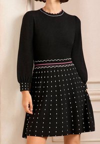 Černé šaty s dlouhými nabíranými rukávy, vroubkovaným výstřihem, texturovaným pasem a plisovanou sukní se vzorem bílých čtverců.