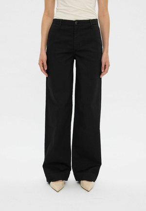 Personne portant un pantalon noir taille haute à jambes larges, associé à des chaussures pointues beiges et un top blanc sans manches rentré.