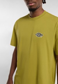Muž v olivově zeleném tričku s krátkým rukávem, na levé části hrudi se nachází černé logo Dickies, na jednobarevném světlém pozadí.