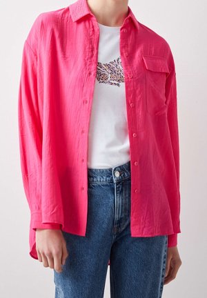 Persona que lleva vaqueros azules, una camiseta blanca con diseño de estampado de leopardo y una camisa de botones rosa brillante abierta con bolsillos en el pecho.