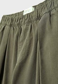 Pantalon taille élastique vert olive avec une étiquette de marque visible et un détail de poche avant.