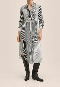 Femme portant une robe midi rayée noir et blanc à rayures verticales avec une ceinture à la taille et des bottes noires jusqu'aux genoux.