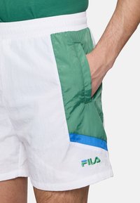 Pantaloni shorts bianchi con un pannello laterale verde e blu, realizzati in materiale leggero, dotati di tasca laterale e logo ricamato.