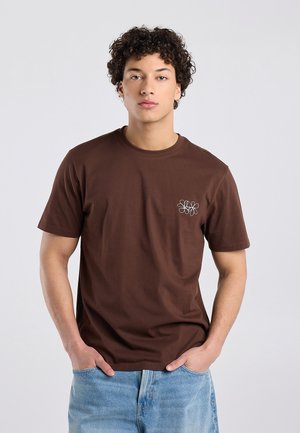 Jonge man met kort, krullend haar draagt een bruin T-shirt met een klein wit bloemenmotief en lichtblauwe spijkerbroek, staat met de handen in de zakken.