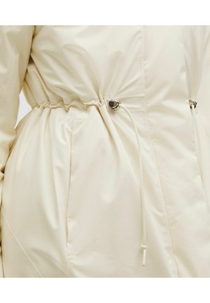 Artimos plano smėlio spalvos švarko juosmuo su reguliuojamu raišteliu ir sidabriniu ryšiu, matyti sklandus audinys ir šoninės kišenės siūlė.