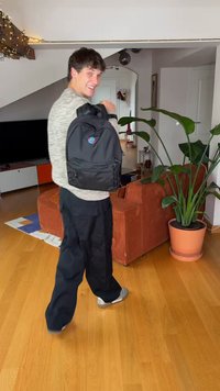 Černý batoh s logem, kapsami na zip a bočními síťovanými držáky. Nosený přes rameno, doplněný lehkým svetrem a tmavými kalhotami.