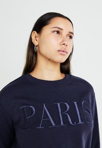 Sweatshirt azul-marinho com "PARIS" bordado numa tonalidade mais clara. Apresenta um decote redondo e punhos canelados. O modelo usa brincos de argola prateados.
