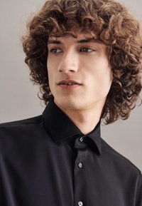 Camicia nera con colletto strutturato, tessuto liscio e bottoni lucidi; indossata da un modello con capelli ricci su uno sfondo neutro.