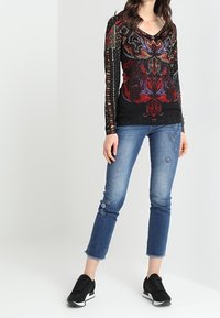 Haut à manches longues noir à motifs avec des accents rouges et verts, associé à un jean bleu clair orné de broderies florales et des baskets noires.