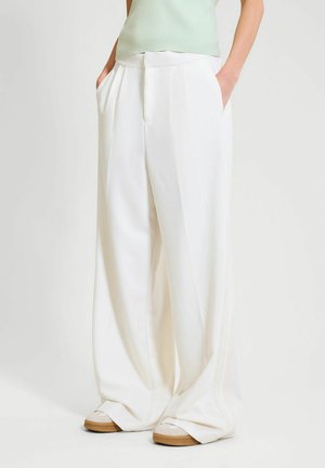 Mujer con pantalones blancos de pierna ancha y tiro alto con bolsillos laterales y zapatillas de color claro, de pie sobre un fondo liso.