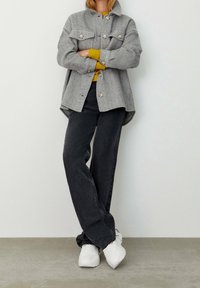 Personne portant une veste grise à boutons sur un pull moutarde, un jean large noir et des baskets blanches, se tenant contre un mur uni.