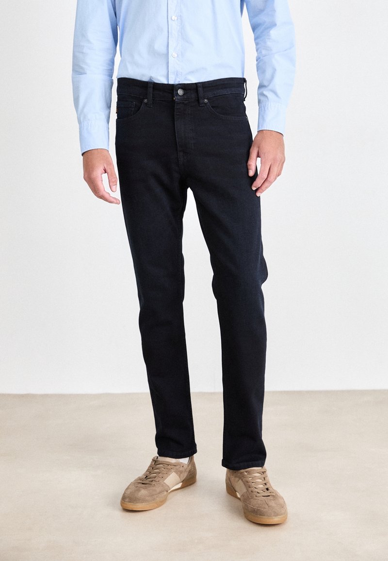 Jean en denim foncé coupe slim avec un design classique à cinq poches et une texture lisse ; porté avec une chemise boutonnée bleu clair.