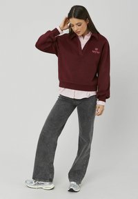 Sweat-shirt bordeaux avec un col en V, des poignets côtelés et un logo brodé, superposé sur une chemise rayée rose, associé à un jean large gris.
