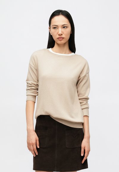 Pull beige clair à manches longues, col rond, et poignets retroussés, associé à une jupe en daim marron foncé avec deux poches avant.