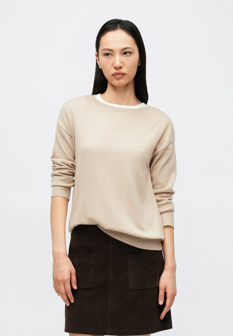 Gaiši bēša džemperis ar garām piedurknēm, apaļu kaklu un uzlocītām aprocēm, kombinēts ar tumši brūnu zamšādas svārku ar divām priekšējām kabatām.