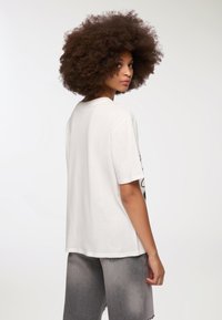 Biała bawełniana koszulka z krótkimi rękawami, okrągłym dekoltem i luźnym krojem, pokazana z tyłu. Połączona z szarymi dżinsowymi szortami.