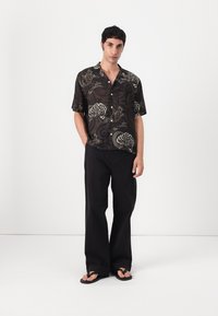 Black short-sleeve shirt with swirling white and gold floral patterns, worn with loose black trousers and flip-flops. Minimalistic design.