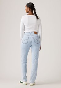 Γυναίκα με πλεγμένα μαλλιά, φοράει λευκό τοπ με μακριά μανίκια, ανοιχτό μπλε τζιν με ψηλή μέση και λευκά αθλητικά με πράσινες λεπτομέρειες, όρθια γυρισμένη προς τα πίσω.