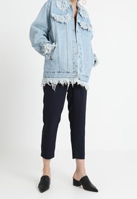 Giacca di jeans azzurro chiaro con bordi sfrangiati e colletto classico, abbinata a pantaloni taper navy e mule nere a punta.