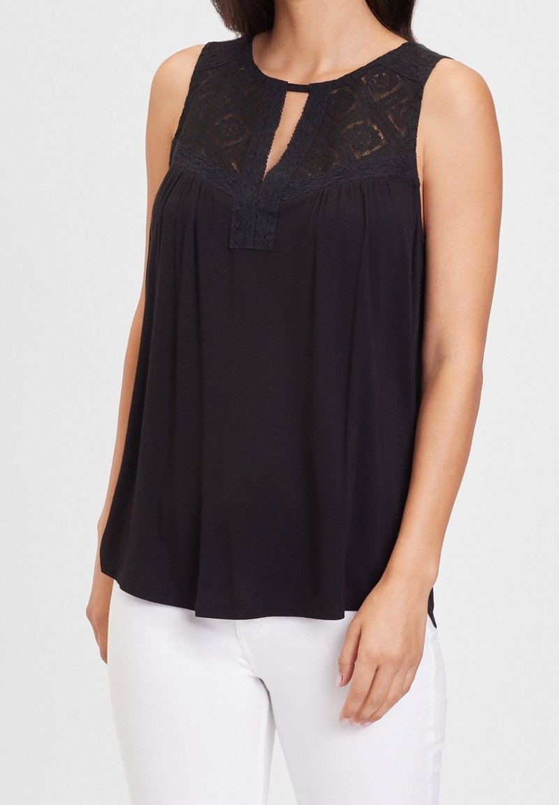 Blouse noire sans manches avec empiècement en dentelle et encolure trou de serrure, portée avec un pantalon blanc, montrée des épaules aux hanches.