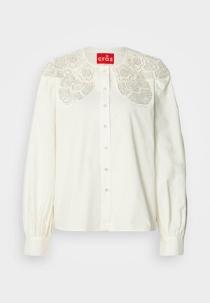 Chemise blanche boutonnée avec col en dentelle brodée décorative. Présente des manches longues avec poignets froncés et une coupe décontractée.