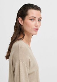 Beige pletený svetr s kulatým výstřihem a dlouhými rukávy. Měkká textura a uvolněný střih, zobrazený na modelce z boku.