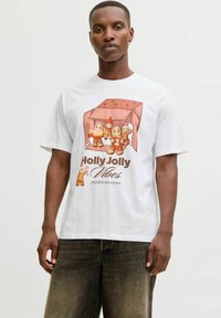 Bărbat îmbrăcat cu un tricou alb decorat cu personaje festive din turtă dulce și textul "Holly Jolly Vibes Fresh & Delicious", combinat cu blugi negri decolorati.
