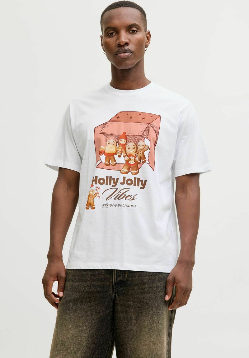 Bărbat îmbrăcat cu un tricou alb decorat cu personaje festive din turtă dulce și textul "Holly Jolly Vibes Fresh & Delicious", combinat cu blugi negri decolorati.