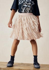 Jasnoróżowa spódnica z falbanami z tiulu o fakturowanym wyglądzie, z dopasowanym pasem i warstwową konstrukcją, w połączeniu z ciemnymi skarpetkami i butami.