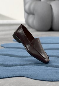 Chaussure en cuir marron à enfiler avec une finition lisse, présentant un motif embossé crocodile et un petit talon, posée sur un tapis bleu texturé.
