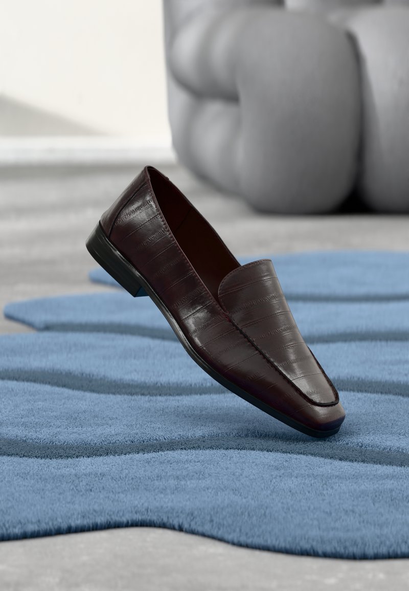 Chaussure en cuir marron à enfiler avec une finition lisse, présentant un motif embossé crocodile et un petit talon, posée sur un tapis bleu texturé.