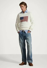 Mężczyzna w białym swetrze z wzorem amerykańskiej flagi, z jasnoniebieską koszulą na spodzie, niebieskich dżinsach i brązowych butach, stojący z rękami w kieszeniach.