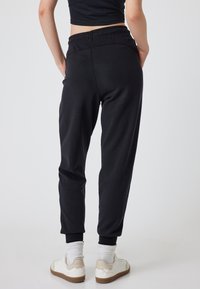 Pantalones de chándal negros con cintura y puños elásticos, hechos de una tela suave. Llevados con calcetines blancos y zapatillas deportivas. Sin patrones notables.