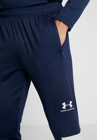 Marine sportbroek met elastische tailleband, zijzakken en wit Under Armour-logo. Gladde textuur, taps toelopend ontwerp en details rond de enkels.