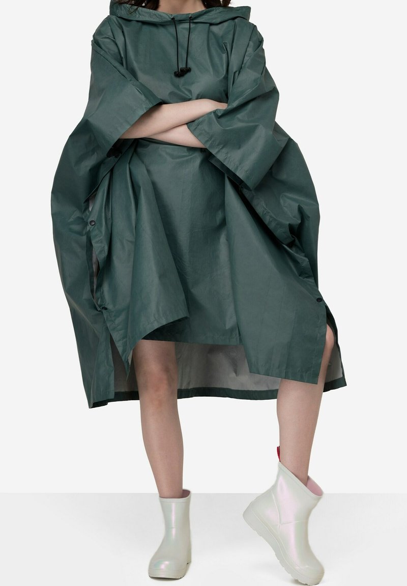 Grøn, oversized vandtæt poncho med hætte, der har snor og sidesnapper. Parret med hvide iriserende ankelstøvler.