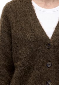 Cardigan brun duveteux avec de gros boutons noirs porté sur un t-shirt blanc côtelé à col rond, visible de la poitrine au cou.