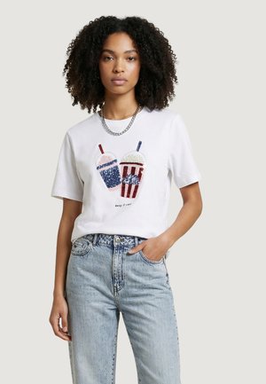 Giovane donna con capelli ricci che indossa una t-shirt bianca con disegni di bevande in paillettes blu e rosse e jeans a vita alta di colore azzurro chiaro.