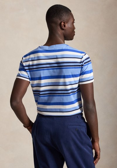 Homme de dos portant une chemise rayée bleue et blanche à manches courtes et un pantalon bleu foncé sur un fond neutre.